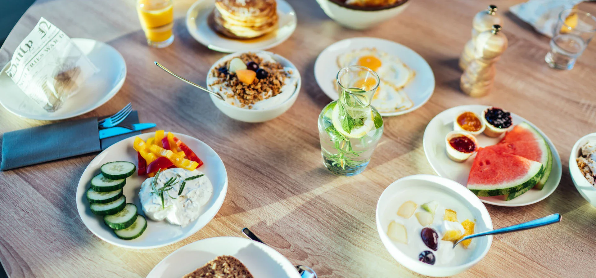 Ein hölzerner Frühstückstisch mit vielen Optionen, darunter frische Früchte wie Wassermelone, Spiegeleier oder Jogurt mit Müsli.