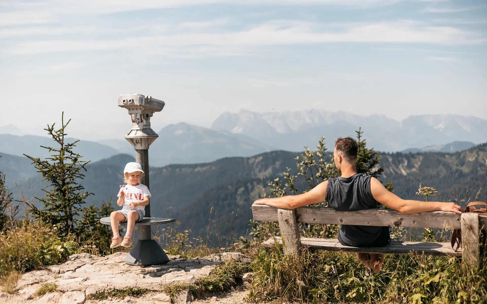 Brückentage Ein Mann sitzt auf einer Bank in den Bergen und schaut zu einem kleinen Mädchen hinüber, das auf der Plattform eines Fernglases sitzt und lächelt. Im Hintergrund erstreckt sich eine Berglandschaft und die Sonne scheint warm.
