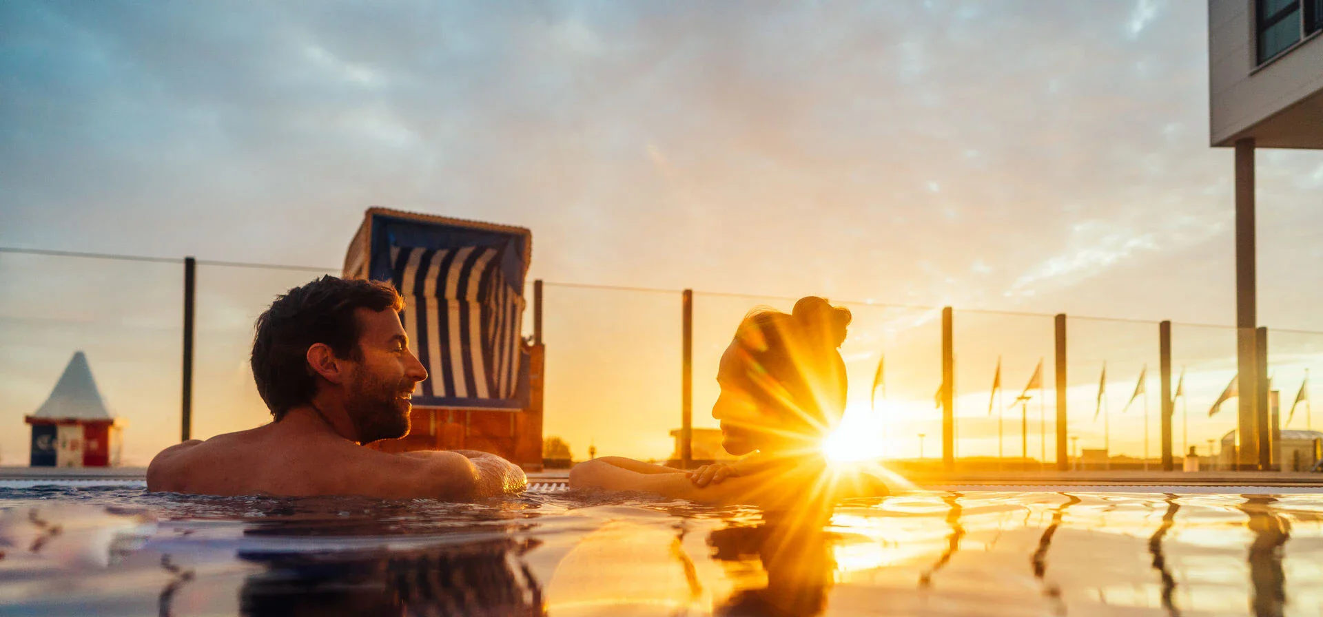Ein Mann und eine Frau schwimmen glücklich bei Sonnenuntergang in einem Pool neben dem ein Strandkorb steht, Die Sonne glitzert golden auf der Wasseroberfläche.