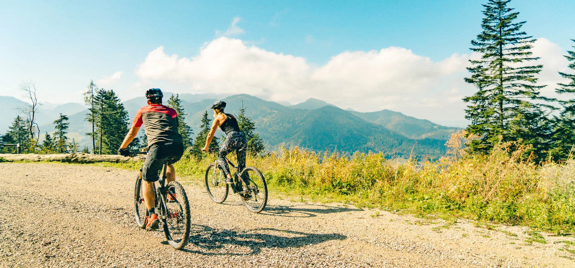 Ein Mann und eine Frau fahren auf Mountainbikes einen Kieselweg in den bergen entlang. Im Hintergrund erstreckt sich eine Berglandschaft und die Sonne scheint.