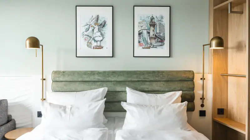 Neues Doppelzimmer im aja Warnemünde Ein Bett mit weißen Kissen und einem grünen Kopfteil, flankiert von zwei Lampen.