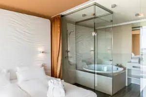 Suite im aja Warnemünde Ein Badezimmer mit einer Glasdusche, Badewanne und Glaswand, davor das Bett.