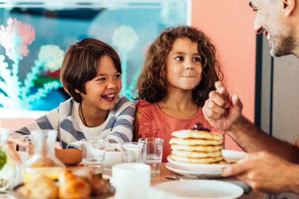 Zwei Kinder sitzen mit dem Vater im Familienbereich an einem Tisch mit Pfannkuchen, Getränken und Brötchen.