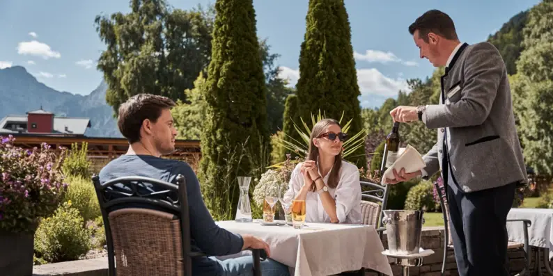 Ein Mann schenkt an einem Tisch im Freien, an dem Personen sitzen, Wein in eine Flasche.