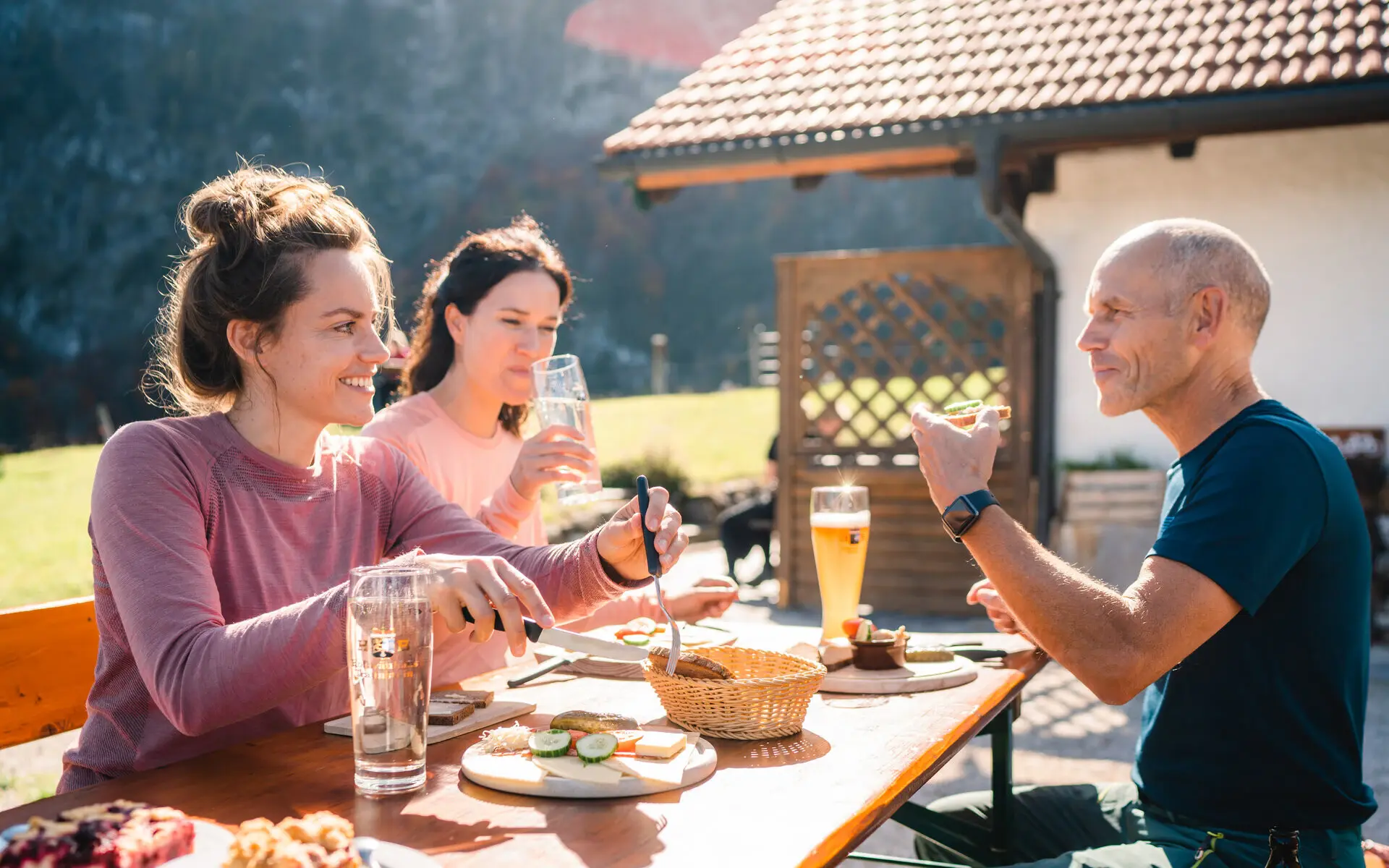 Eine Gruppe an Menschen sitzt an einem Tisch vor einer Hütte mit kühlen Getränken, Brot mit Aufschnitt und. Kuchen. Die Sonne strahlt und im Hintergrund sind die Berge zu sehen. 