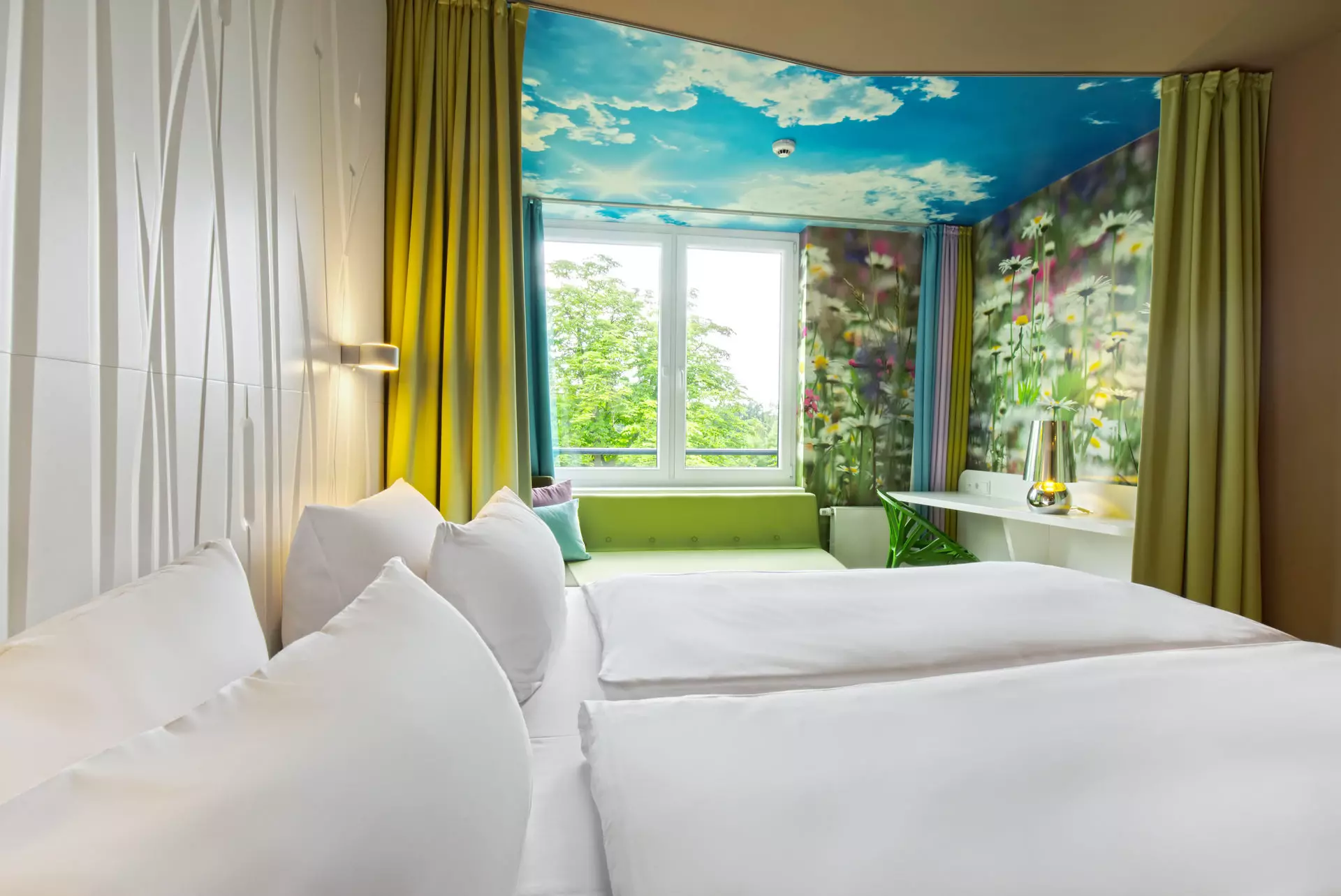 Blick über ein mit weißer Bettwäsche bezogenes Doppelbett vor einer weißen Rückwand mit Struktur.  Im Hintergrund ist ein von blau-grünen und mit Blumenmuster verzierten Wänden umrahmtes Fenster mit Blick ins Grüne zu erkennen. Die Zimmerdecke vor dem Fenster ist mit Himmels-Motiv bedruckt.