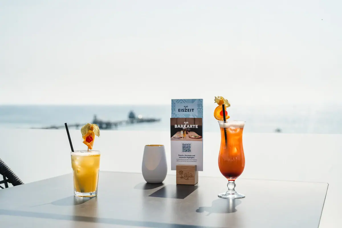 Zwei Cocktails auf einem Tisch in der aja Rooftop Bar Grömitz mit Meerblick und der Seebrücke im Hintergrund.