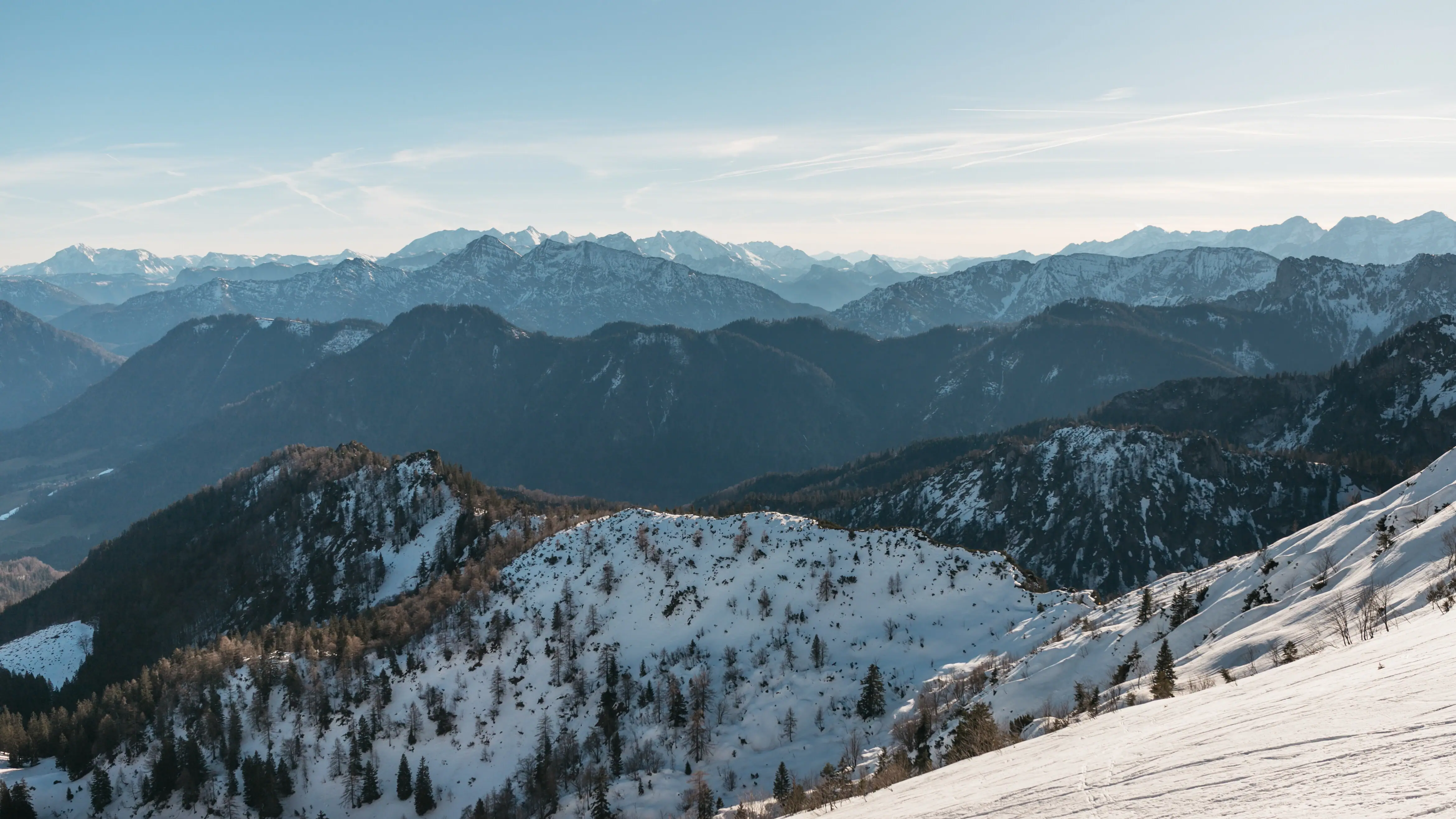 Verschneiter Berg mit Bäumen und weiteren Bergen im Hintergrund.