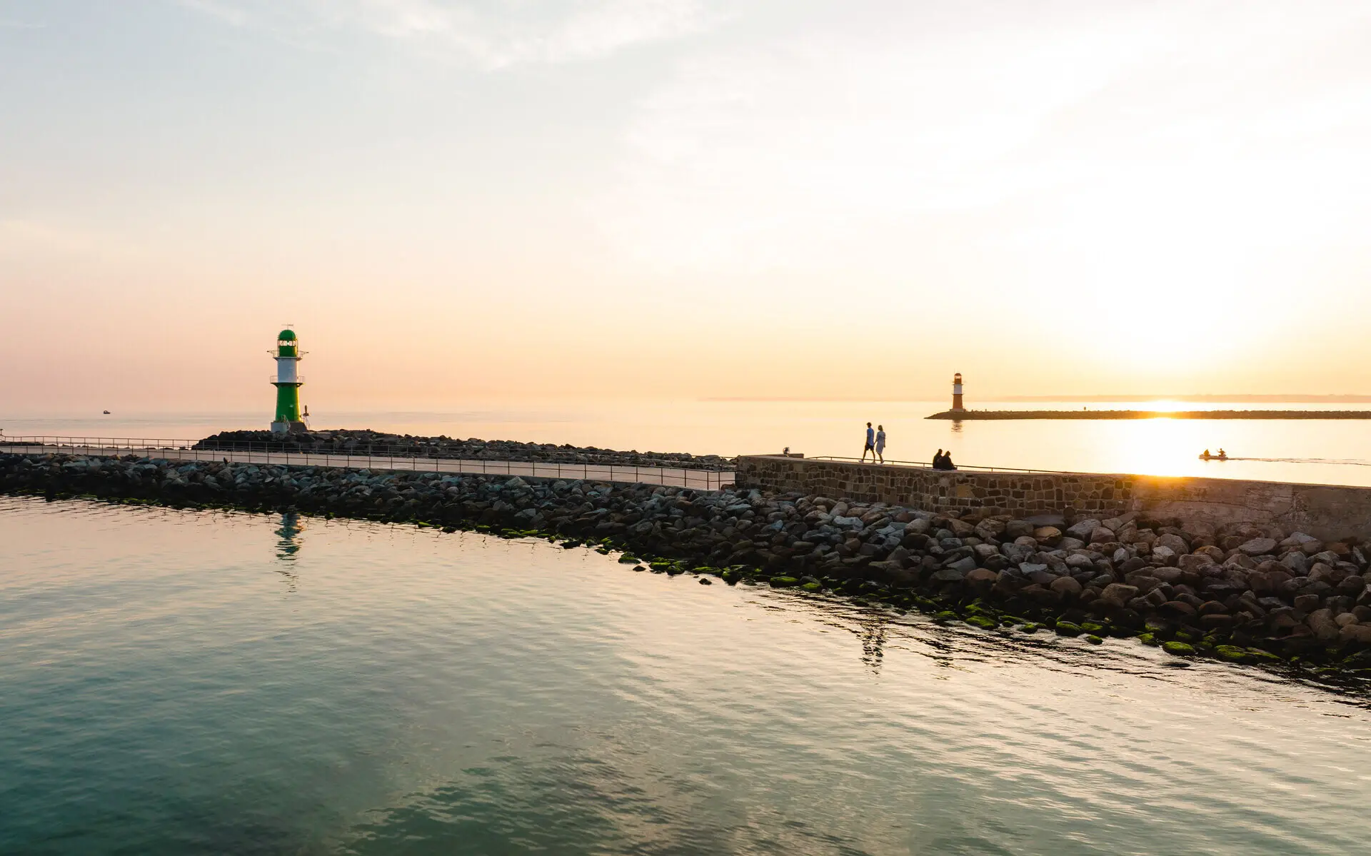 Blick auf ein Gewässer mit einem Leuchtturm in der Ferne bei Sonnenuntergang.