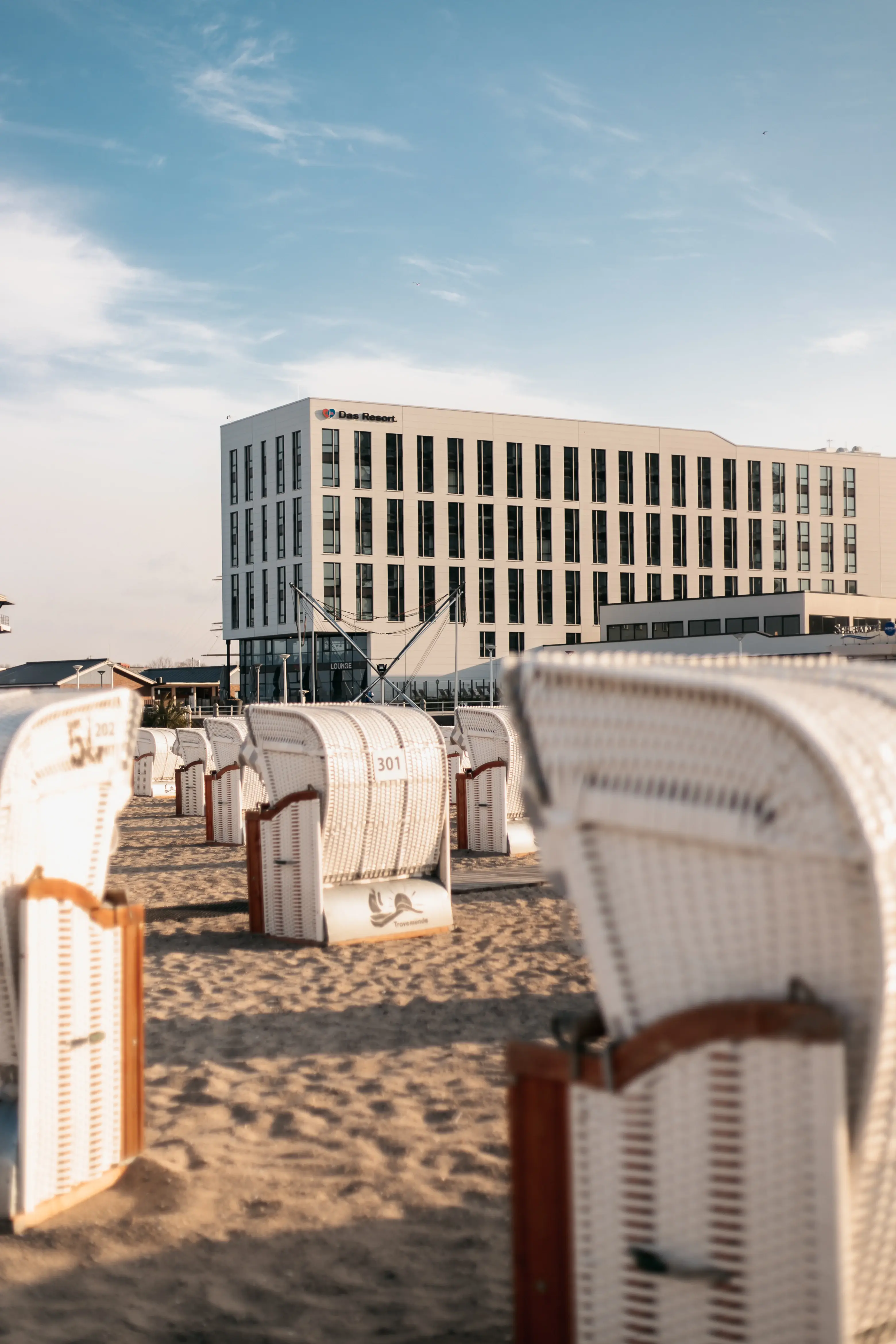 Eine Gruppe von Strandkörben im Sand mit dem aja Hotel Travemünde im Hintergrund.