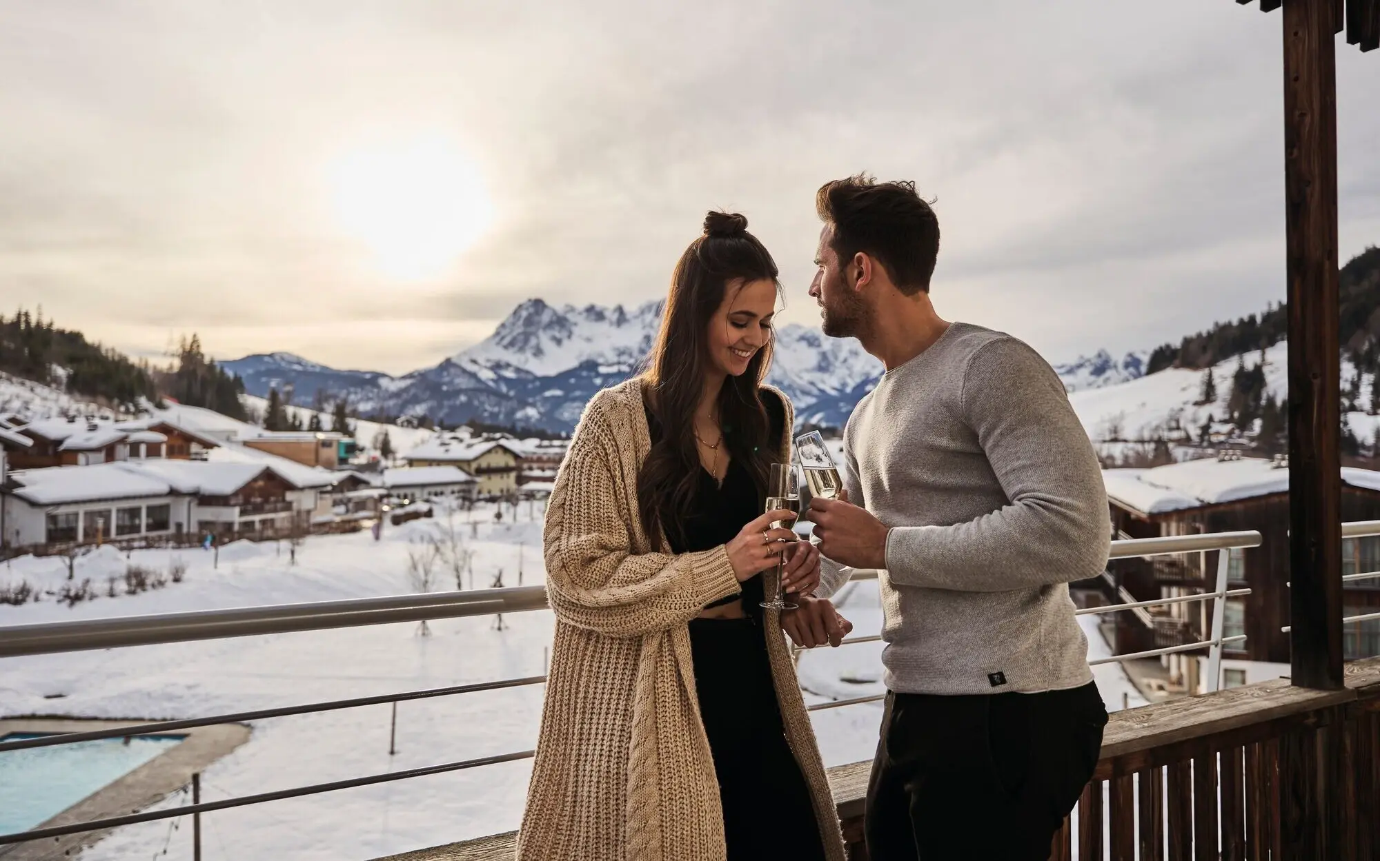 Ein Mann und eine Frau halten auf einem Balkon mit schneebedeckten Bergen im Hintergrund Sektgläser.