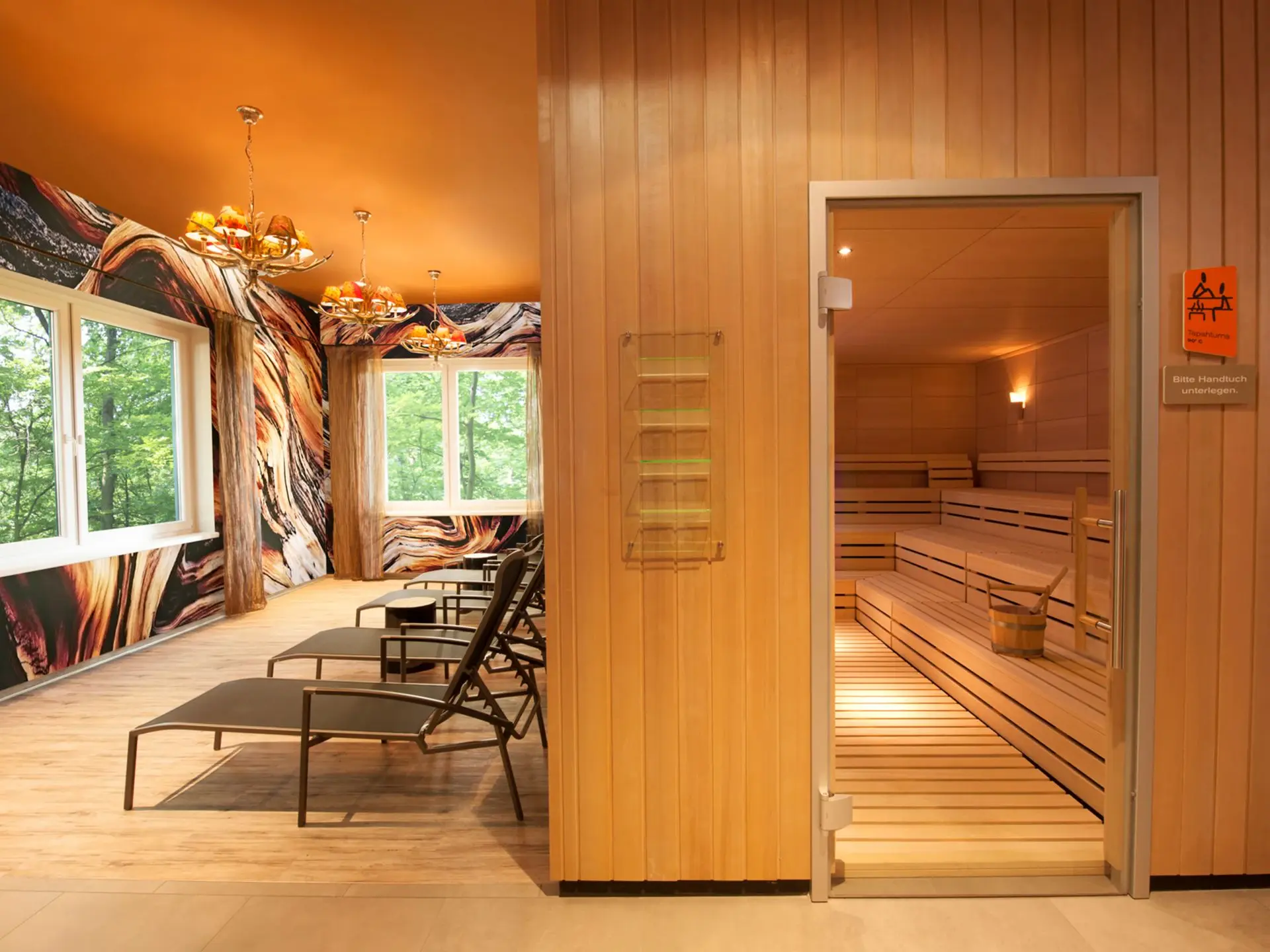 Im rechten Teil ist die Eingangstür zu einer hölzernen Sauna eingelassen in eine mit Holz verkleidete Wand zu sehen. Am linken Rand stehen einige Liegen mit Blickrichtung Fenster mit Ausblick ins Grüne aufgereiht.