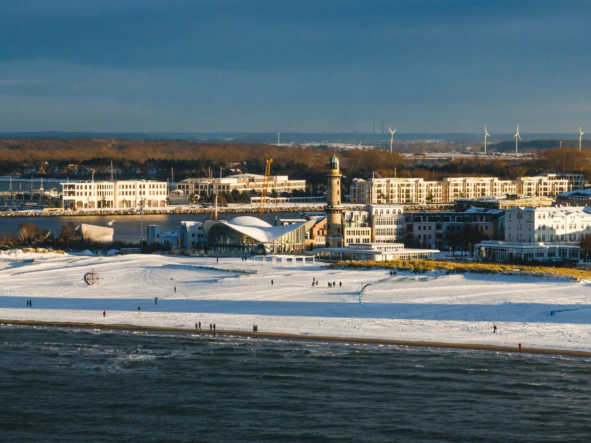 Blick auf Warnemünde im Winter mit schneebedecktem Strand, der Ostsee im Vordergrund und dem markanten Leuchtturm sowie der Teepott-Architektur im Zentrum.