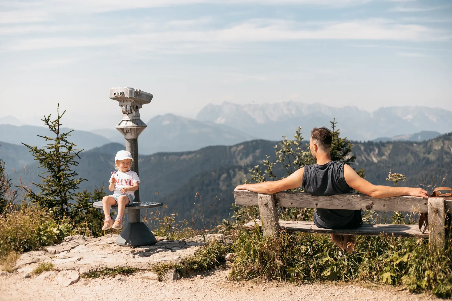 Ein Mädchen sitzt auf dem Fußtritt eines Fernglases und neben ihr ein Mann auf einer Bank, der zu ihr hinüber schaut. Im Hintergrund erstreckt sich eine weite Berg- und Wälderlandschaft und die Sonne scheint warm. 