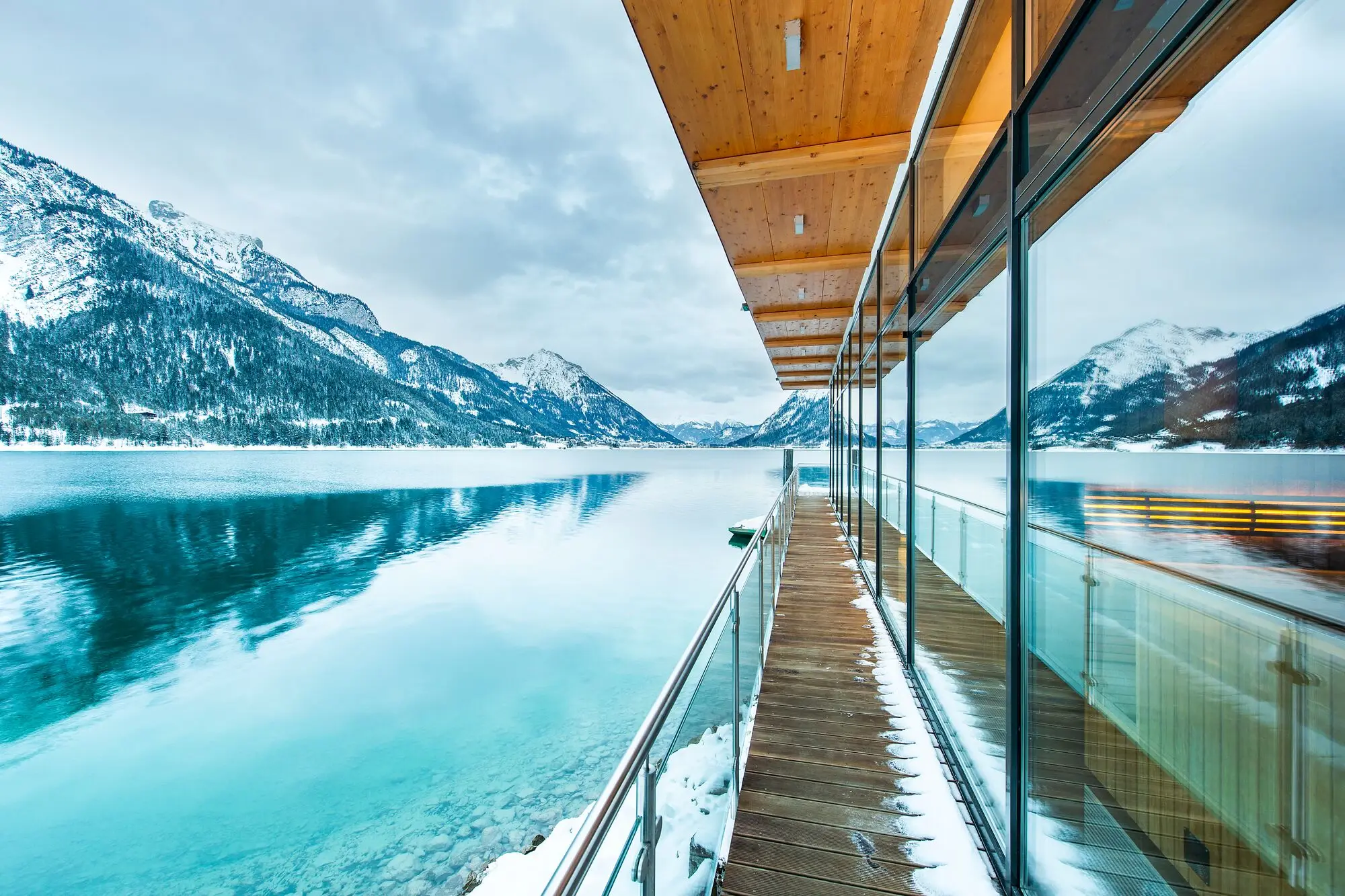 Glaswand mit Holzdeck mit Blick auf einen See, umgeben von Bergen und Wolken.