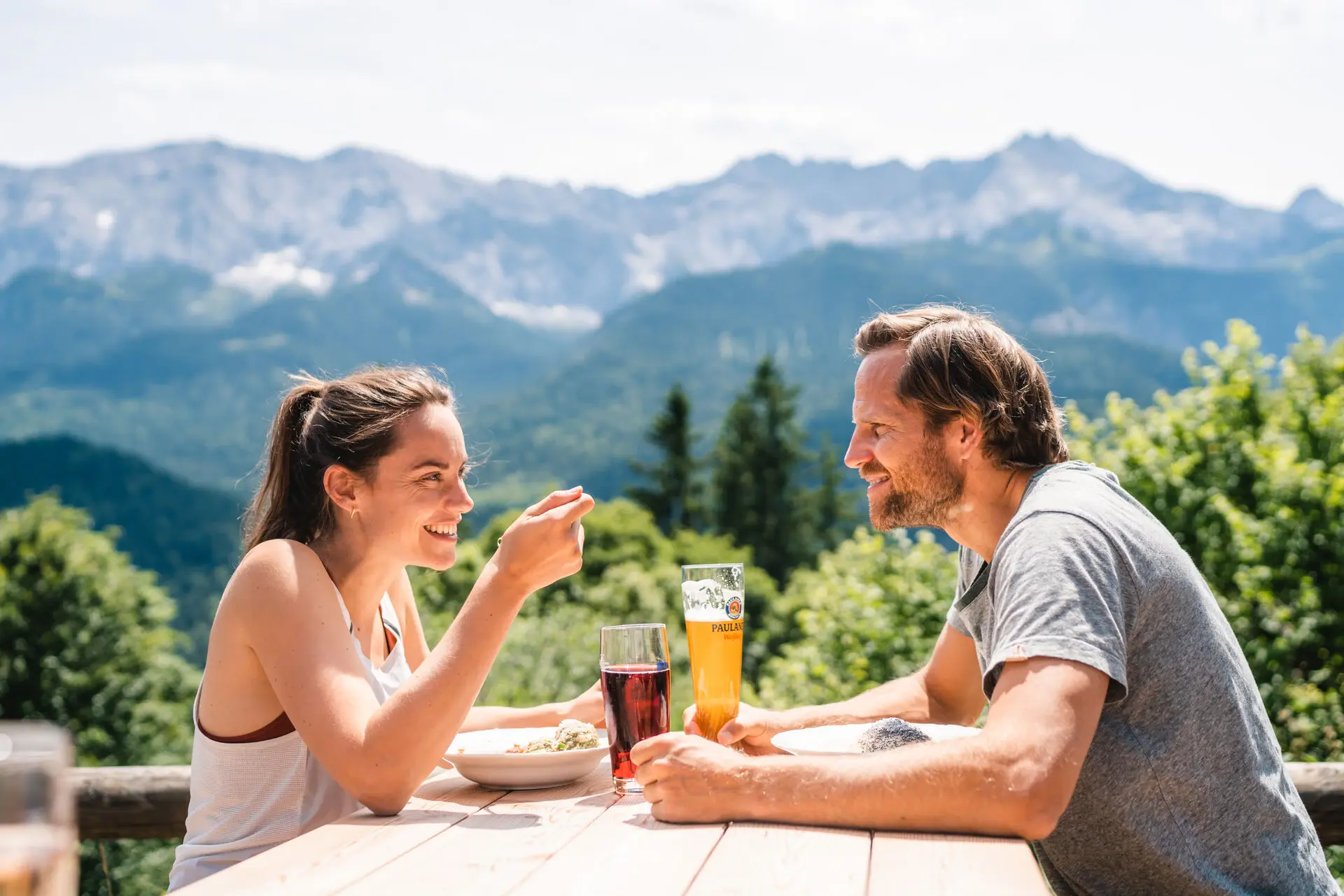 Ein Paar sitzt glücklich an einem Holztisch in den Bergen und genießt Essen und kalte Getränke während der Sommerzeit. 