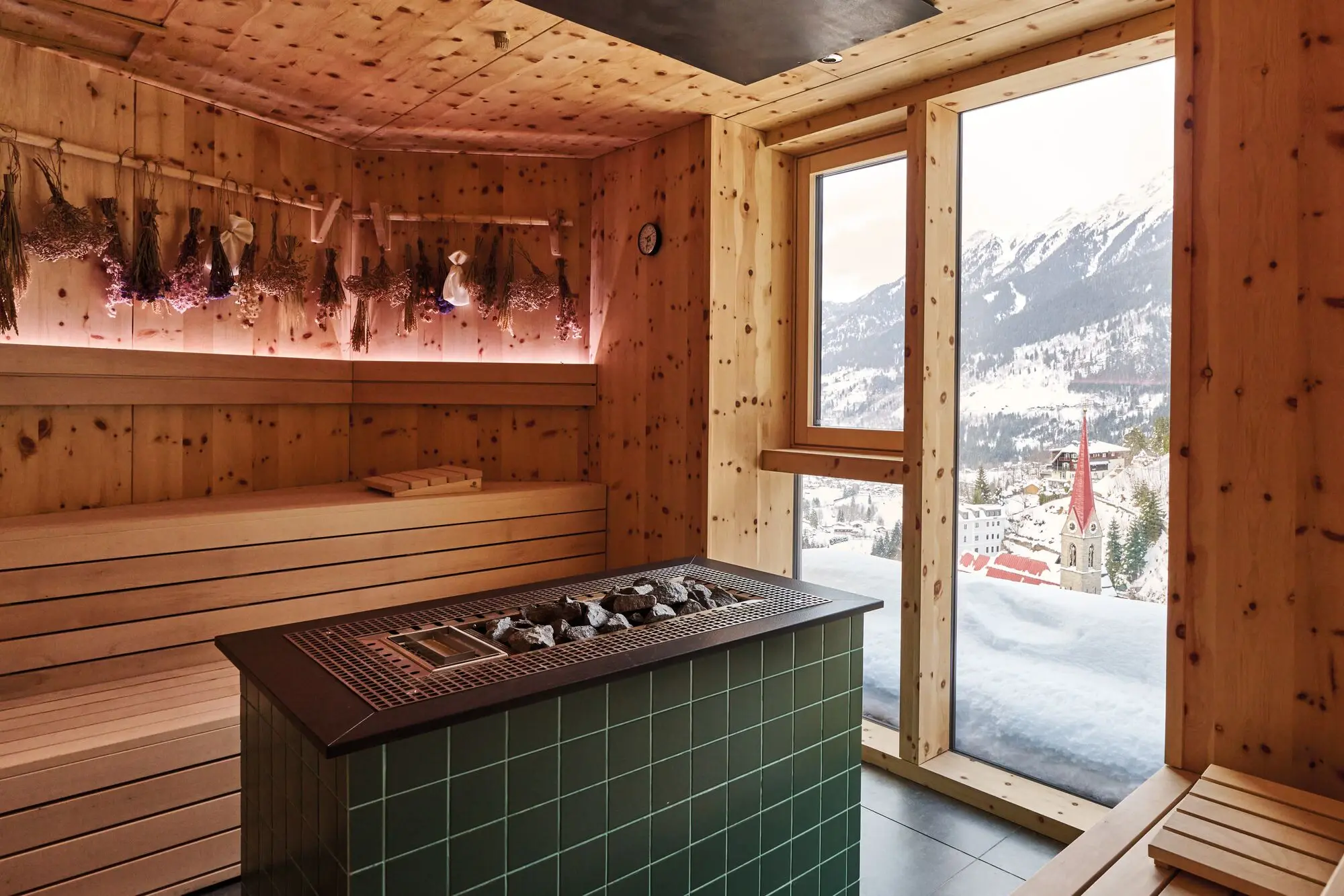 Eine gemütliche Sauna aus Holz mit einem zentralen Saunaofen, dekoriert mit getrockneten Kräuterbündeln an den Wänden. Durch das große Fenster bietet sich ein beeindruckender Blick auf eine verschneite Berglandschaft und eine malerische Kirche im Tal.