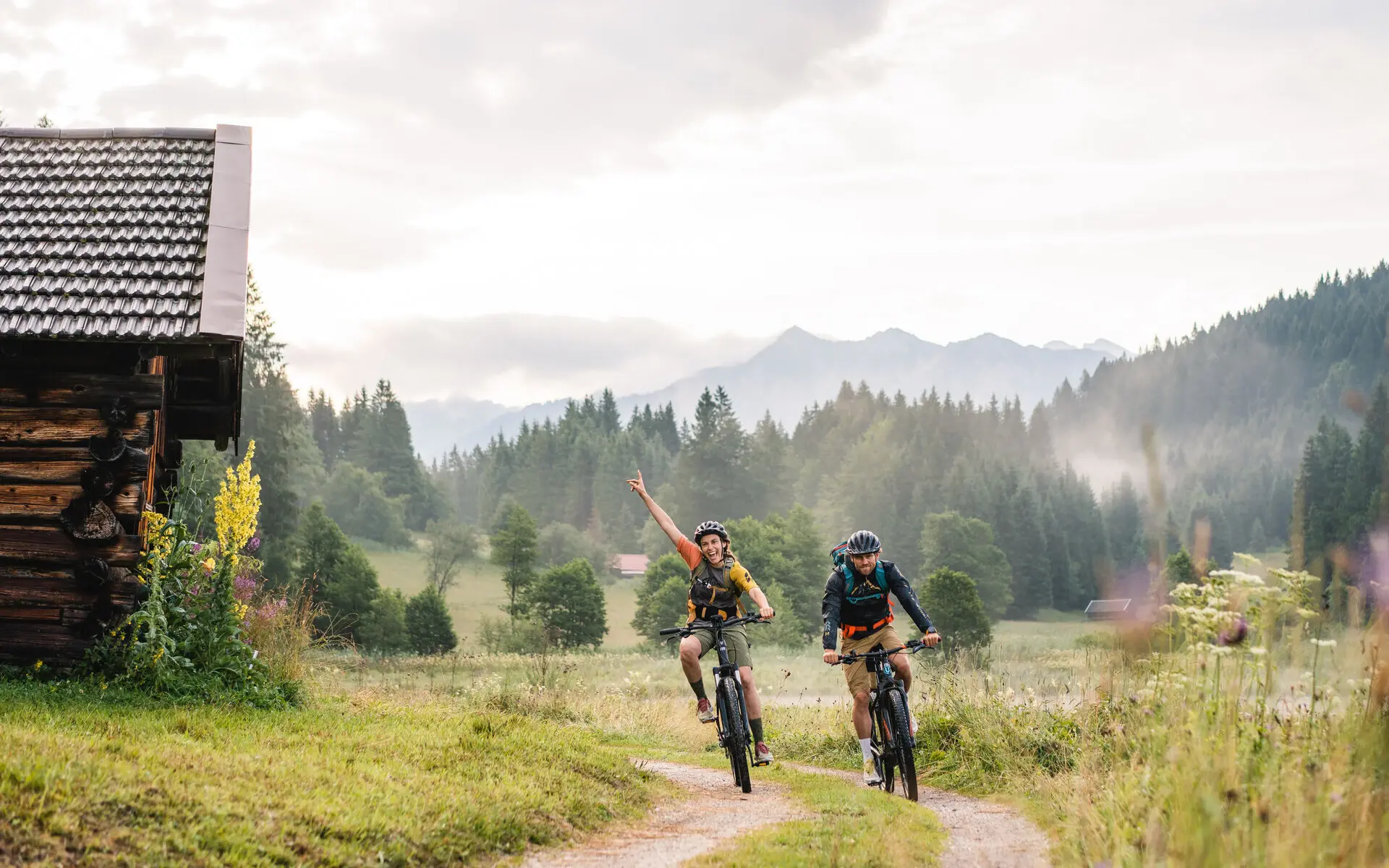 Ein Paar fährt auf Mountainbikes durch die Wälder. Die grüne Landschaft ist von Morgendunst bedeckt und der Himmel leicht grau. 