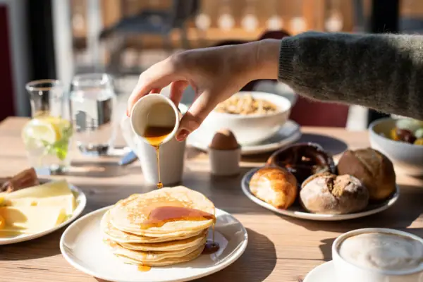 Frühstückstisch im aja Ruhpolding Eine Person gießt Sirup auf einen Stapel Pfannkuchen.