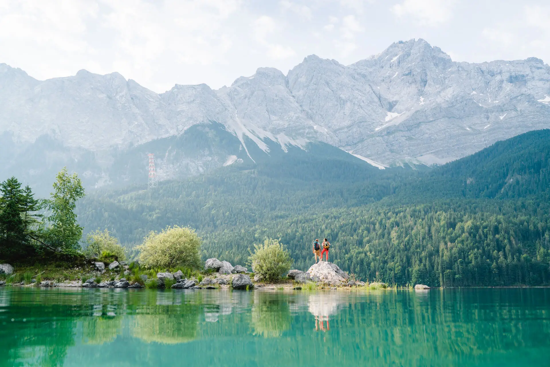 Ein Paar steht auf Steinen an einem grün blauen See und schaut über die dahinter liegenden, tannenbewachsenen Berge in die Weiten. 