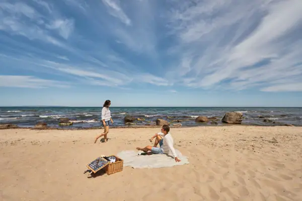 Paar beim Picknick am naturbelassenen Ostseestrand; der Mann sitzt auf einer Decke im Sand, die Frau spaziert barfuß am Wasser entlang, im Vordergrund ein Picknickkorb, im Hintergrund das Meer mit Felsen und weiter Horizont.