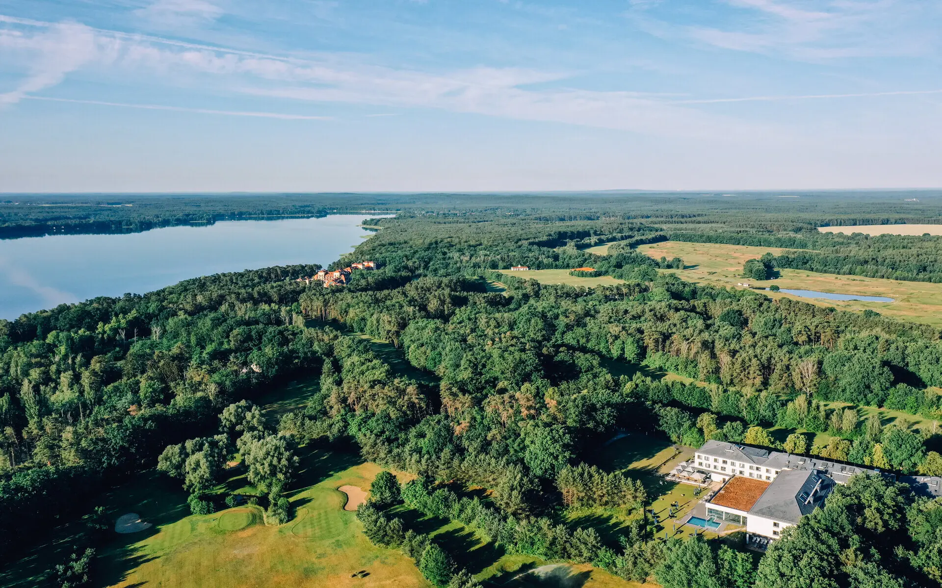 Luftansicht des aja Bad Saarow, umgeben von grüner Natur und einem großen See im Hintergrund. 