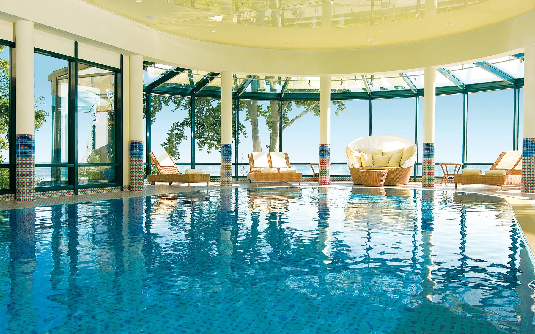 Indoor pool with chairs under a large ceiling, reflecting on the water.