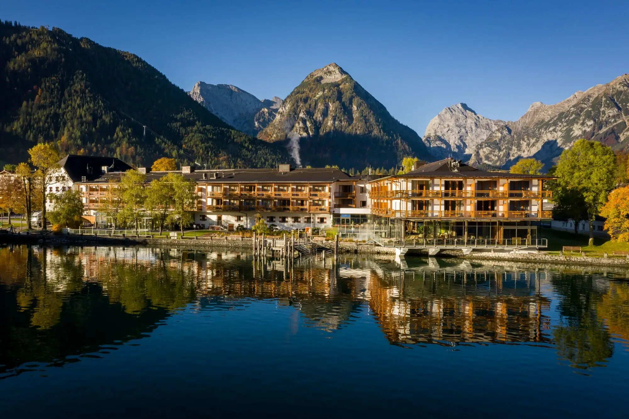aja Achensee Außenansicht vom aja Achensee am See mit Bergen und Bäumen im Hintergrund in warmes Licht getaucht.