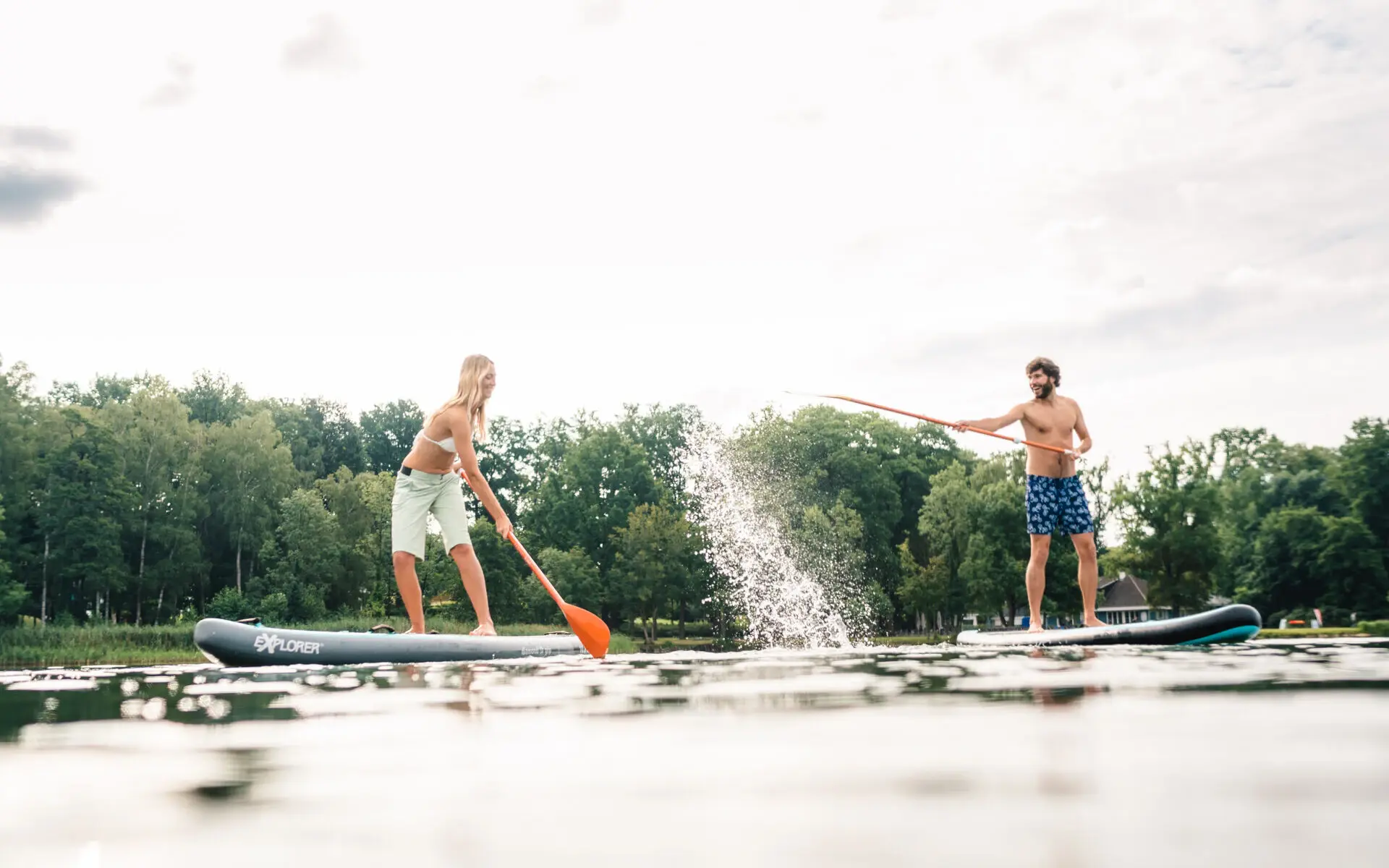 Ein Mann und eine Frau stehen jeweils auf einem Stand-Up Paddling Board und spritzen mit ihren Paddeln Wasser durch die Luft. 