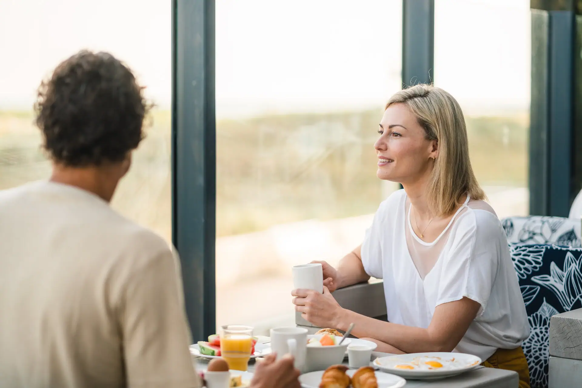 Restaurant im aja Warnemünde Ein Paar sitzt am Frühstückstisch und eine Frau hält eine Tasse und schaut die andere Person an. Im Hintergrund sind die Dünen zu sehen.