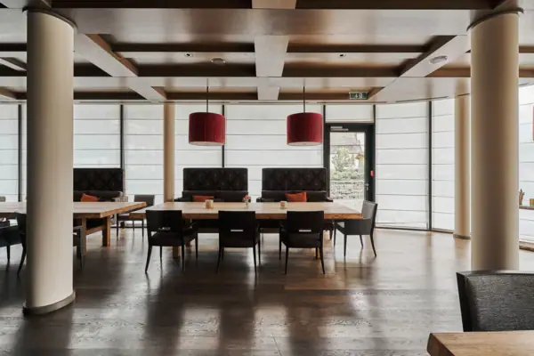 Ein großer Raum mit Holzboden, großen Tischen und roten Lampen. Die Fenster rund um den räum sind mit Sichtschutz verdeckt. 