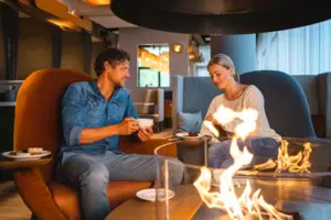 Ein Paar sitzt gemütlich in einer Lounge mit modernen Sesseln und genießt Kaffee und Dessert, während im Vordergrund ein offenes Feuer für eine warme Atmosphäre sorgt.