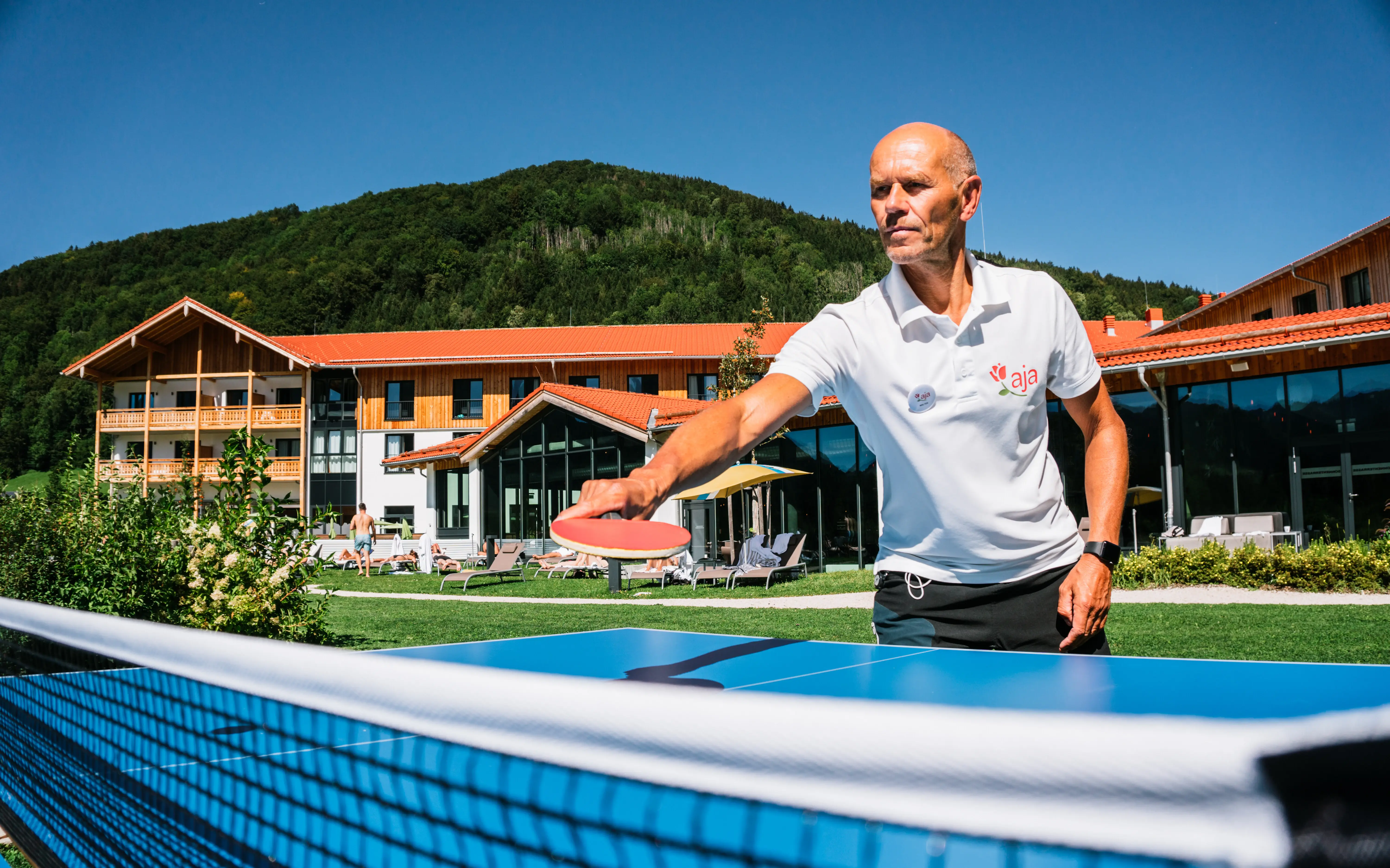 Ein Mann spielt Tischtennis im Außenbereich des aja Resorts vor einer Kulisse aus grünen Hügeln und dem modernen Hotelgebäude mit Holzelementen.