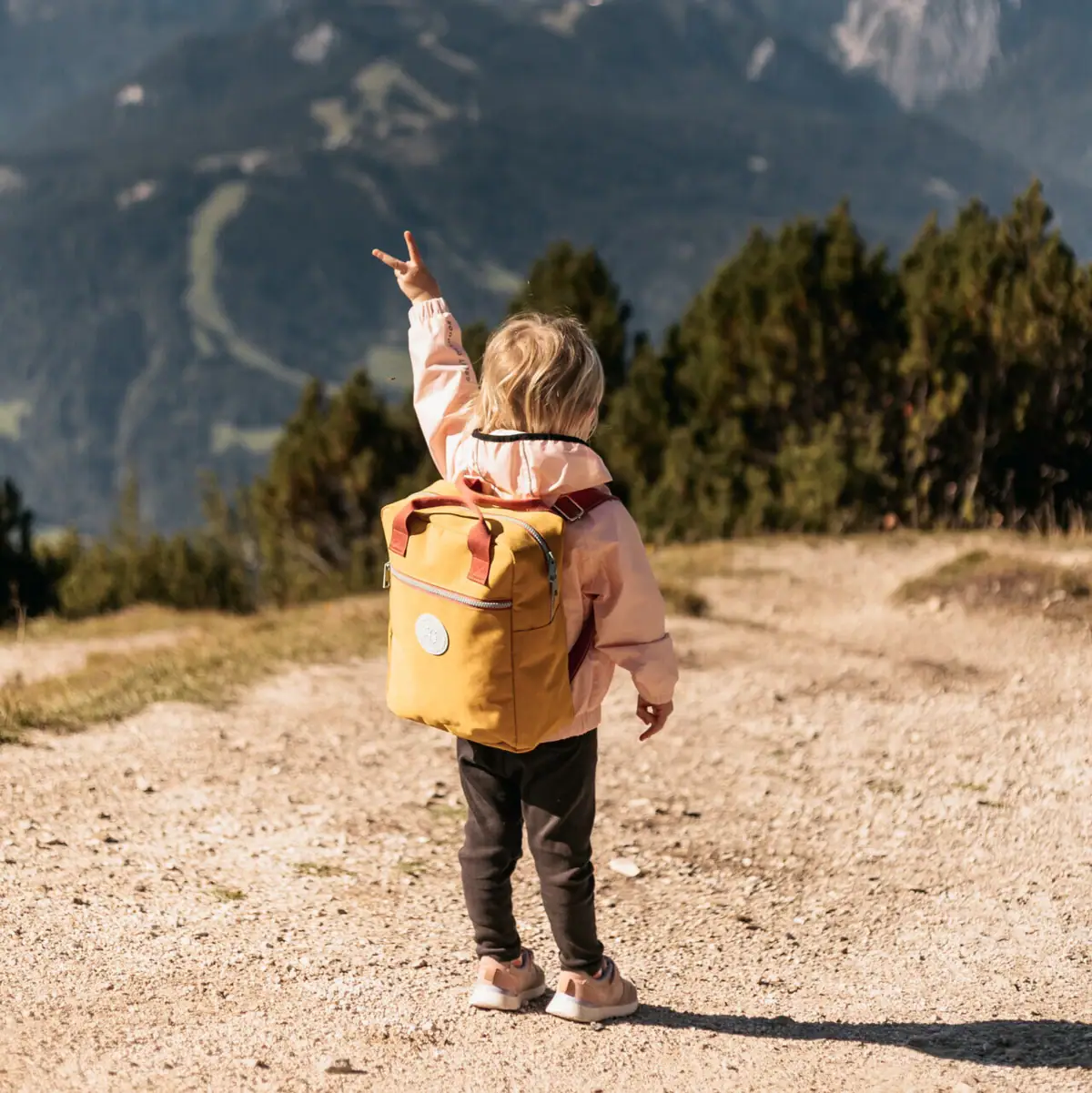 Ein Kind mit gelbem Rucksack steht auf einem Feldweg, im Hintergrund eine Bergkette.