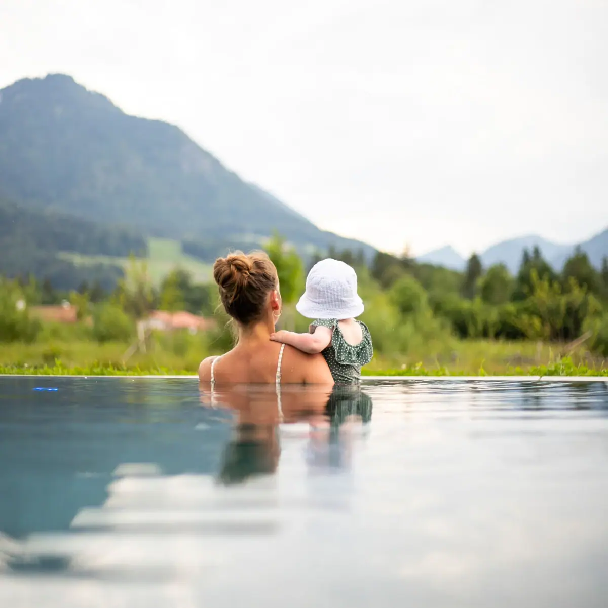 Eine Mutter sitzt mit ihrem Kleinkind im Wasser eines Infinity-Pools und blickt gemeinsam auf eine idyllische Berglandschaft.
