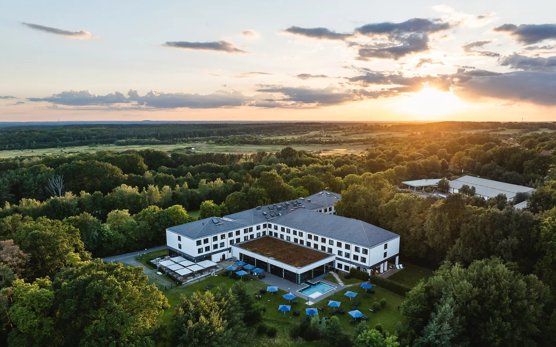 Eine Luftansicht des aja Bad Saarow mit Außenpool in grüner Natur, umgeben von Bäumen bei Sonnenuntergang. 