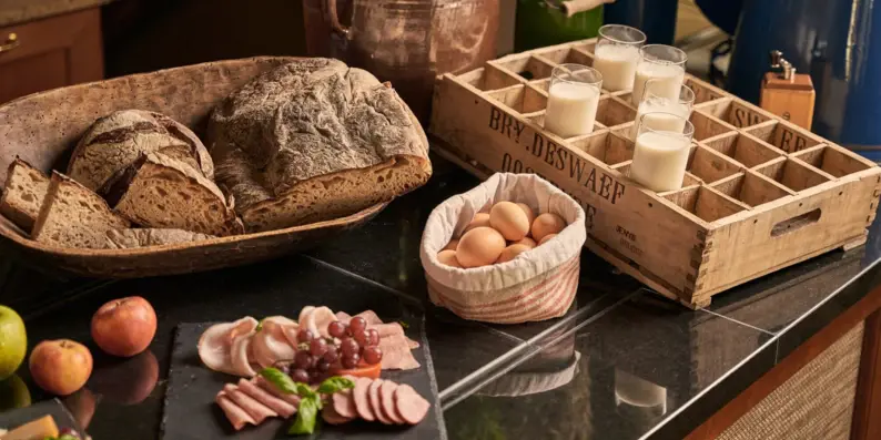 Ein Frühstücksbuffet mit einem großen Brotkorb, einer Wusrplatte, Frühstückseiern, Äpfeln und Gläsern mit Milch.