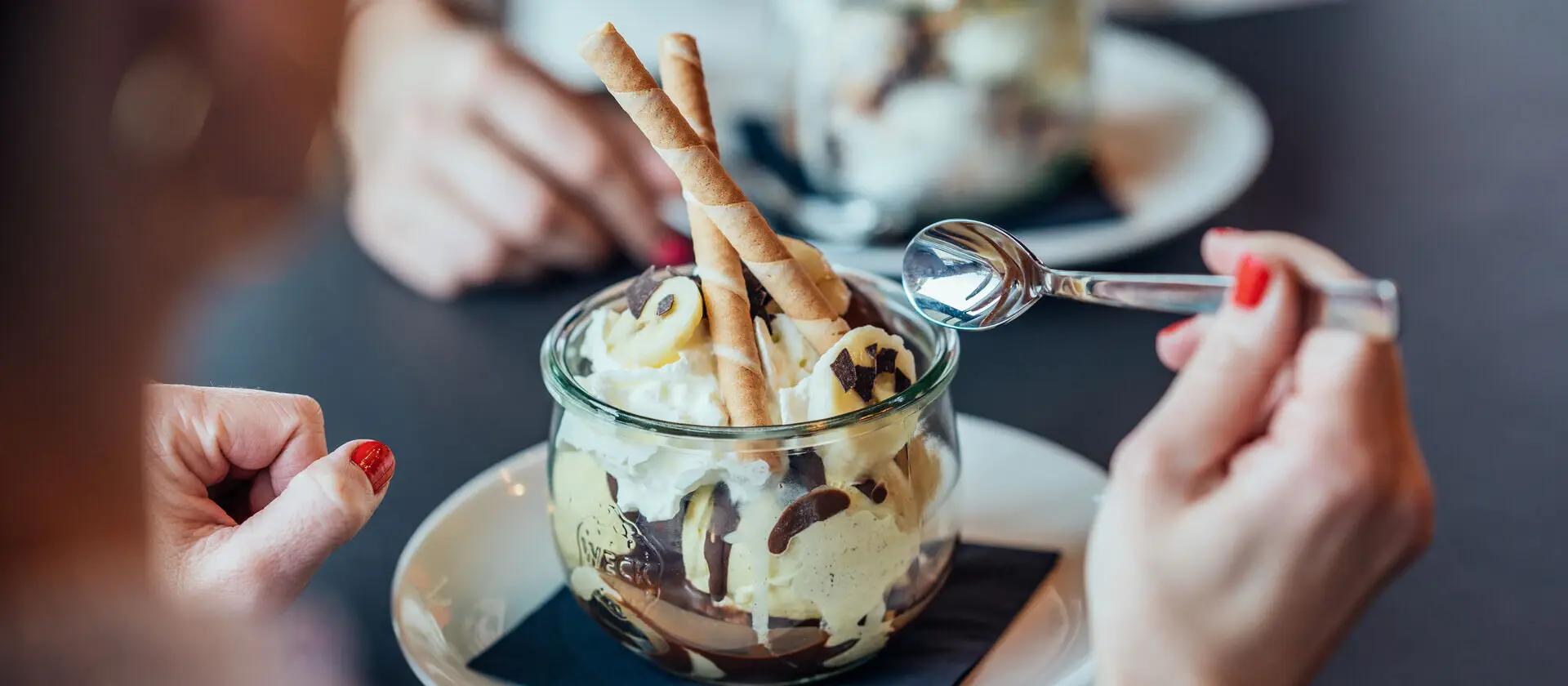 Ein Eisbecher mit Schokolade, Bananenstücken und Sahne in einer Glasschüssel serviert.
