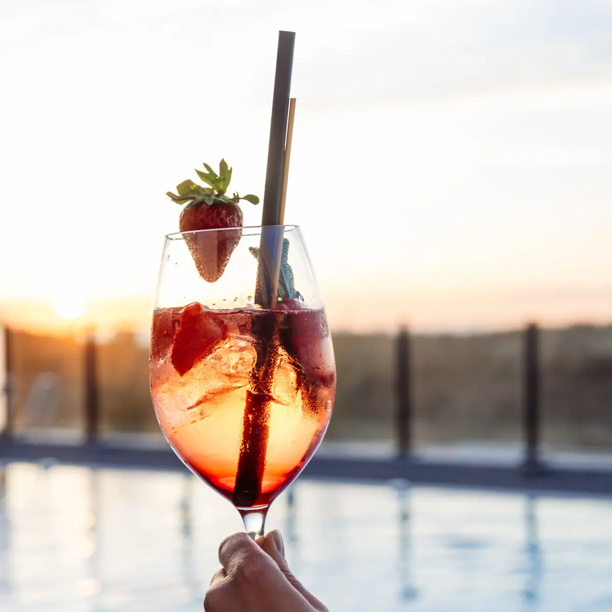 Eine Hand hält ein Getränk mit Erdbeeren, im Hintergrund ist ein Pool zu sehen.