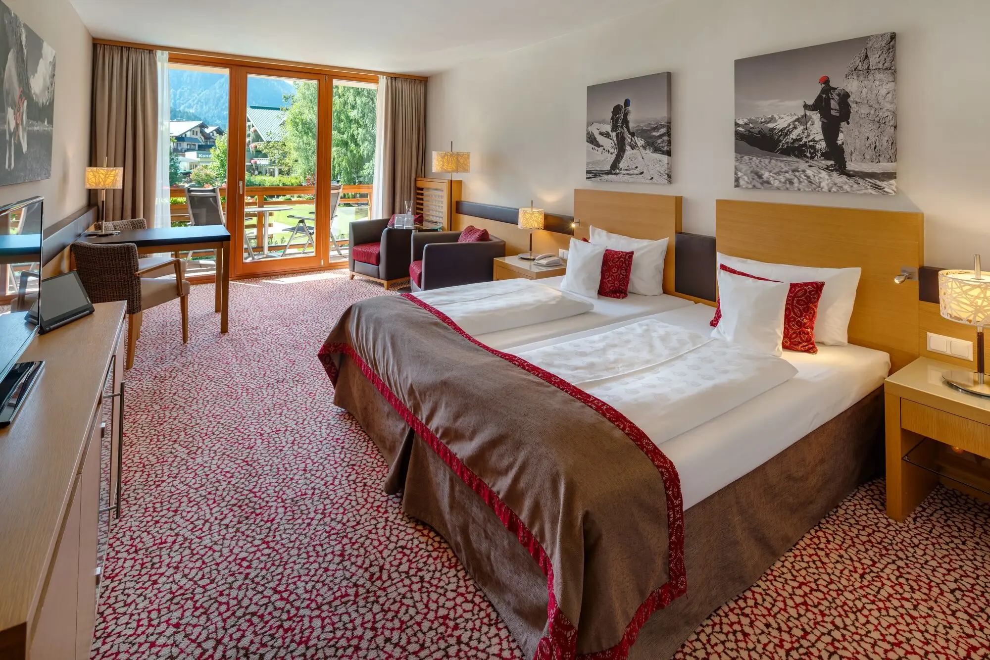 Ein gemütlich eingerichtetes Hotelzimmer mit Doppelbett, roten Kissen und warmem Holzdekor, dekoriert mit Bildern von Adlern und Berglandschaften an der Wand. Neben dem Bett stehen Nachttische mit modernen Lampen, und ein Flachbildfernseher ist an der Wand befestigt