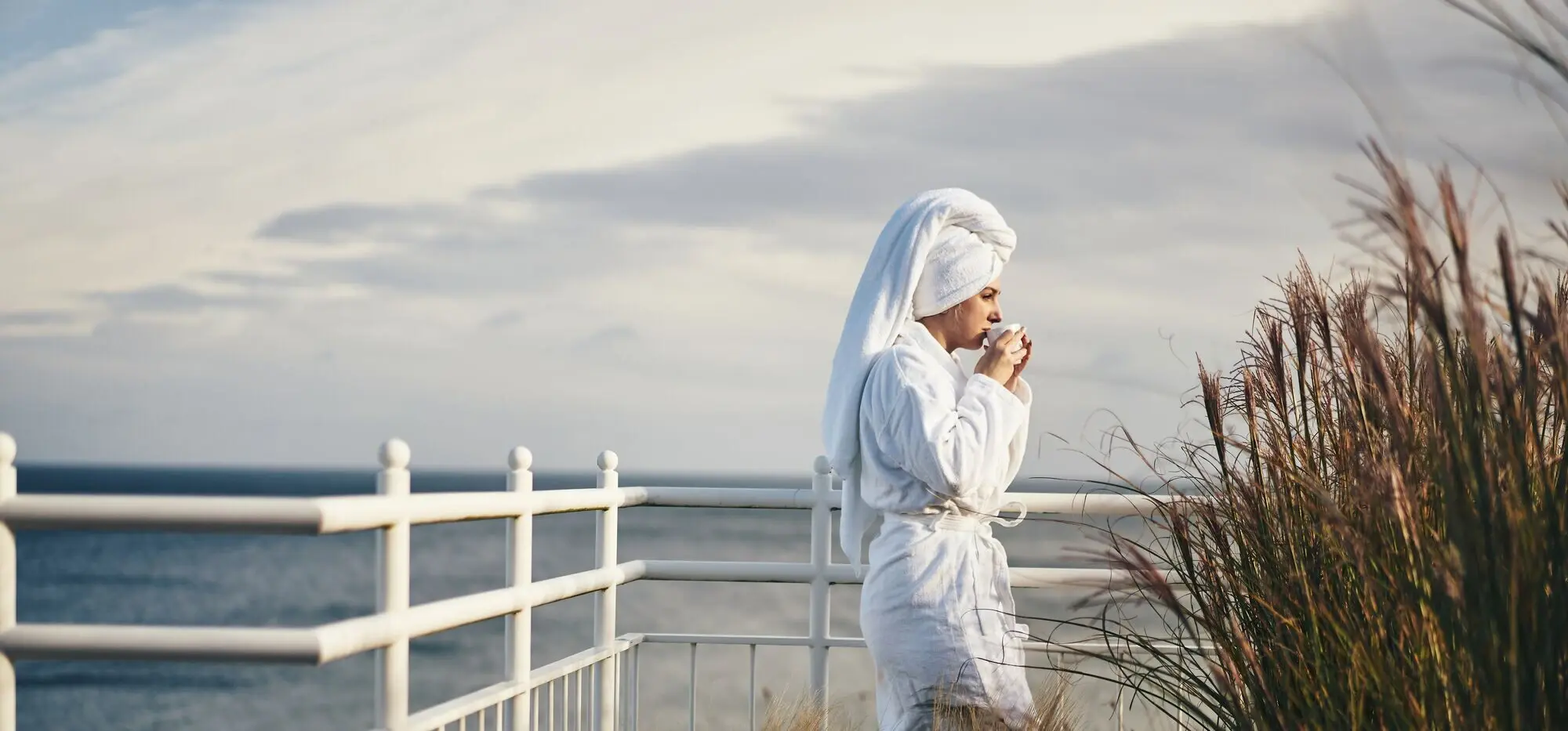 Woman in a bathrobe drinking from a cup outdoors.