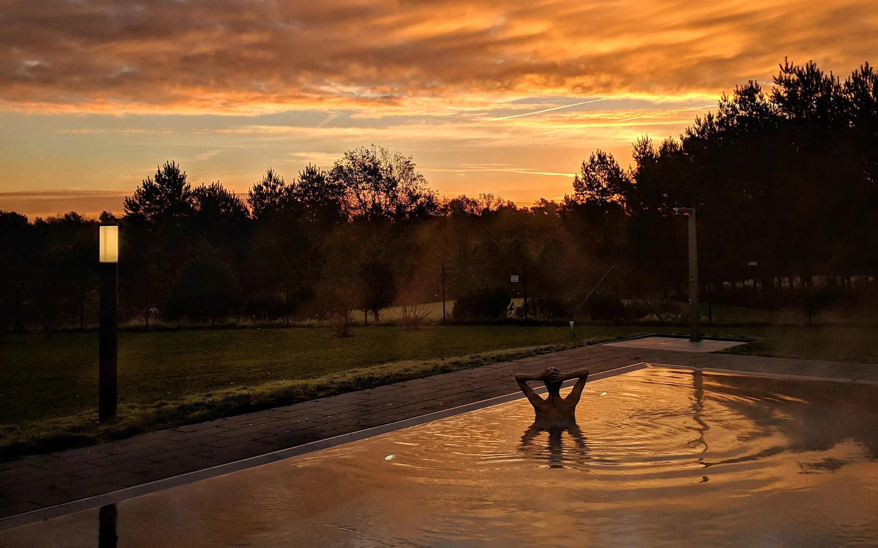 Der Außenpool des aja Bad Saarow in der Abenddämmerung. In der Mitte des Pools sind Umrisse einer Person zu erkennen, welche die Hände hinter dem Kopf verschränkt. Im Hintergrund sind die Schatten von Bäumen zu erkennen, die Wolken am Himmel sind orange-rot gefärbt und tauchen den Außenbereich in ein abendliches Licht.