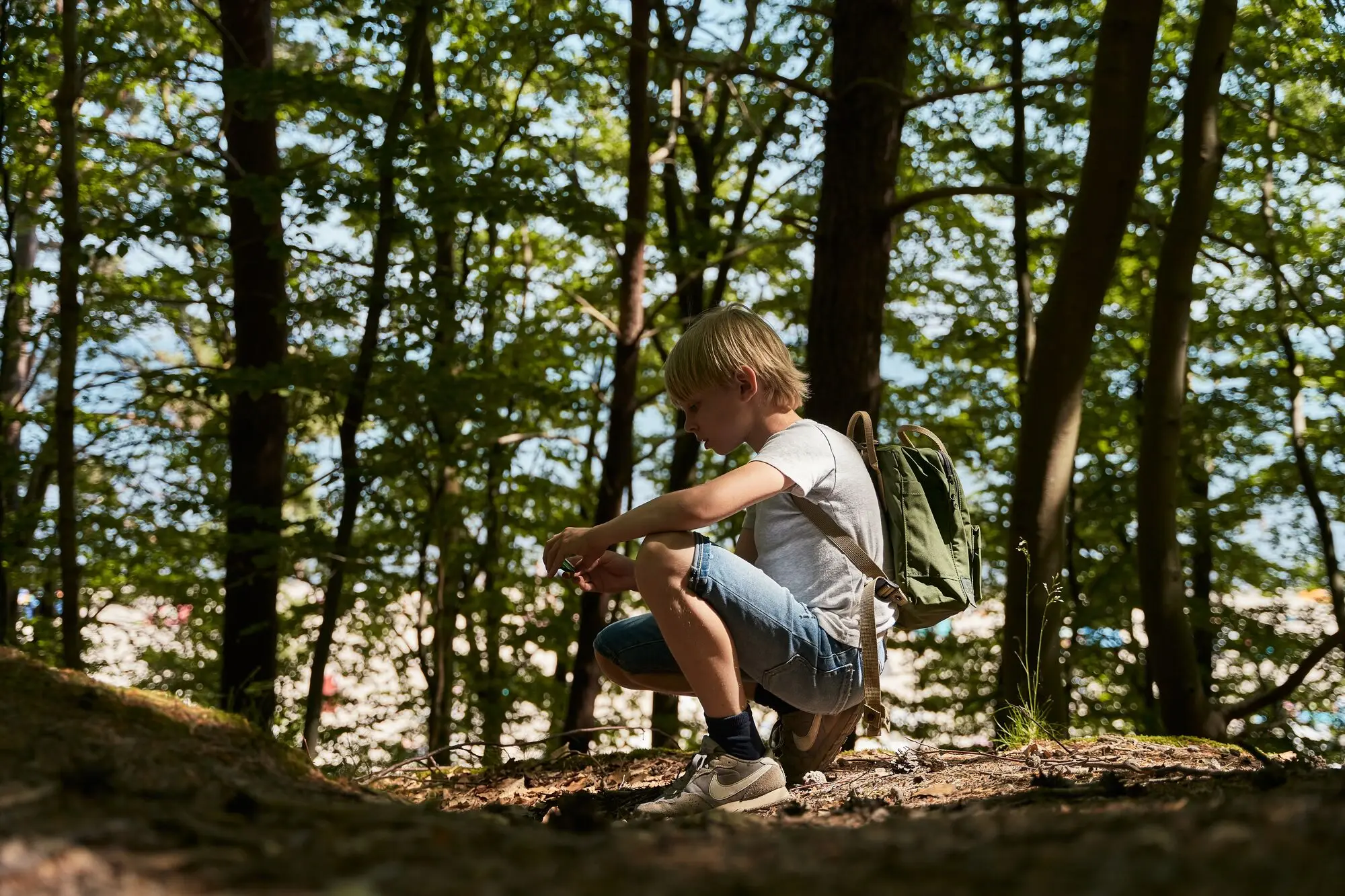 Ein Junge kniet im Wald, umgeben von Bäumen, in Outdoor-Kleidung und Wanderschuhen.