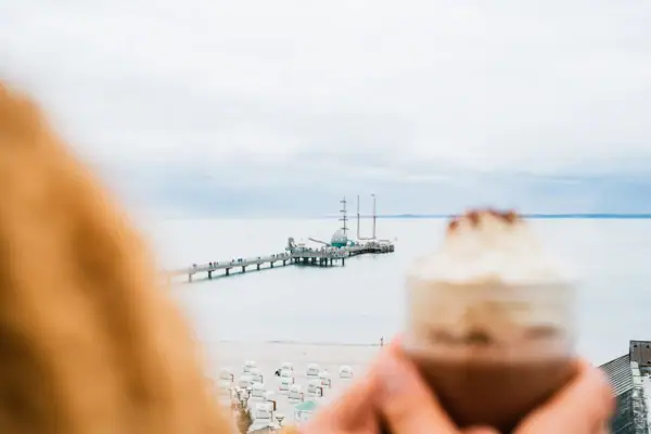 Eine Person hält eine heiße Schokolade mit Sahne und im Hintergrund ist ein Pier zu sehen.
