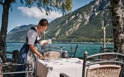 Eine Frau in einem Dirndl deckt einen Tisch an einem See ein.