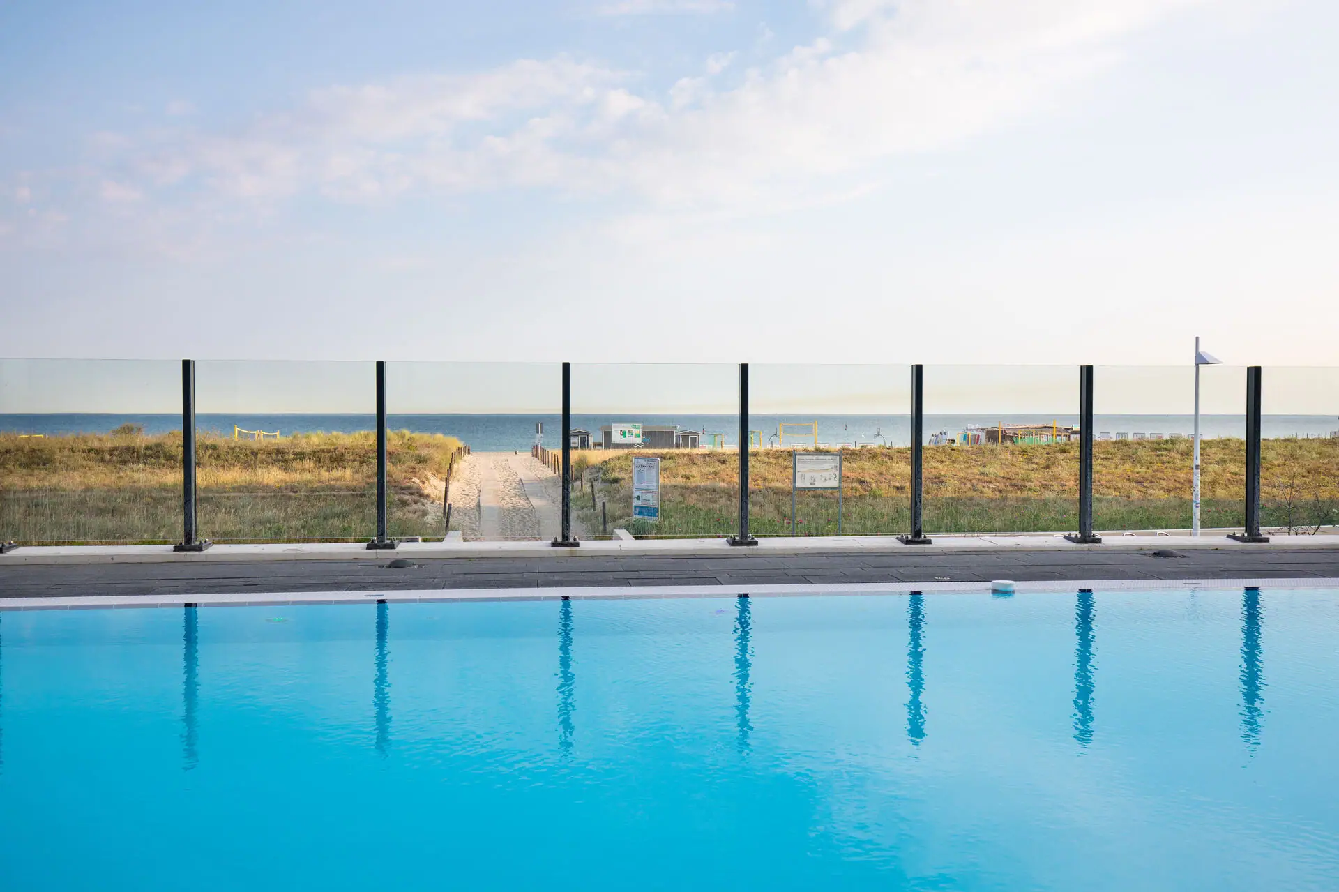 Ein Pool mit Zaun und Rasen im Vordergrund, dahinter ein Strand.