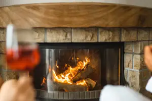 Zwei Personen sitzen in Bademänteln vor einem steinernen Kamin. Das Feuer brennt friedlich und warm, eine Person hält ein Glas mit rötlichem Getränk in der Hand.