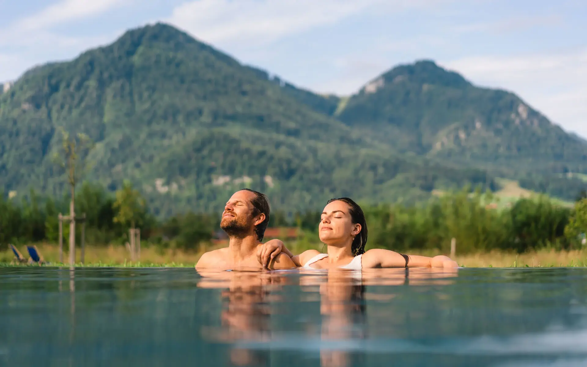 Ein Mann und eine Frau entspannen im Außenpool unter freiem Himmel. Sie stützen sich an den Beckenrand gelehnt nebeneinander auf. Die Augen sind geschlossen und die Gesichtszüge wirken entspannt. Im Hintergrund ist die Berglandschaft der Chiemgauer Alpen zu erkennen.
