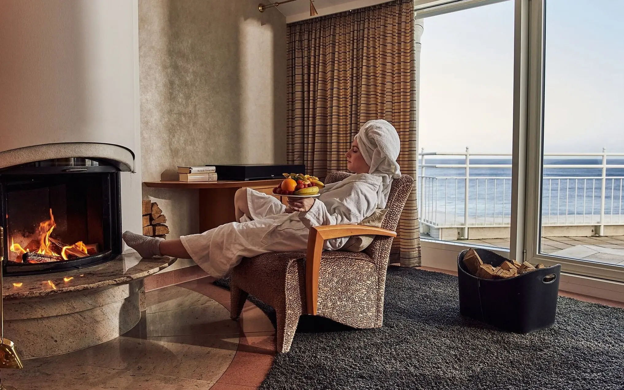 Person in a bathrobe sits on a chair next to a fireplace and a bowl of fruit.