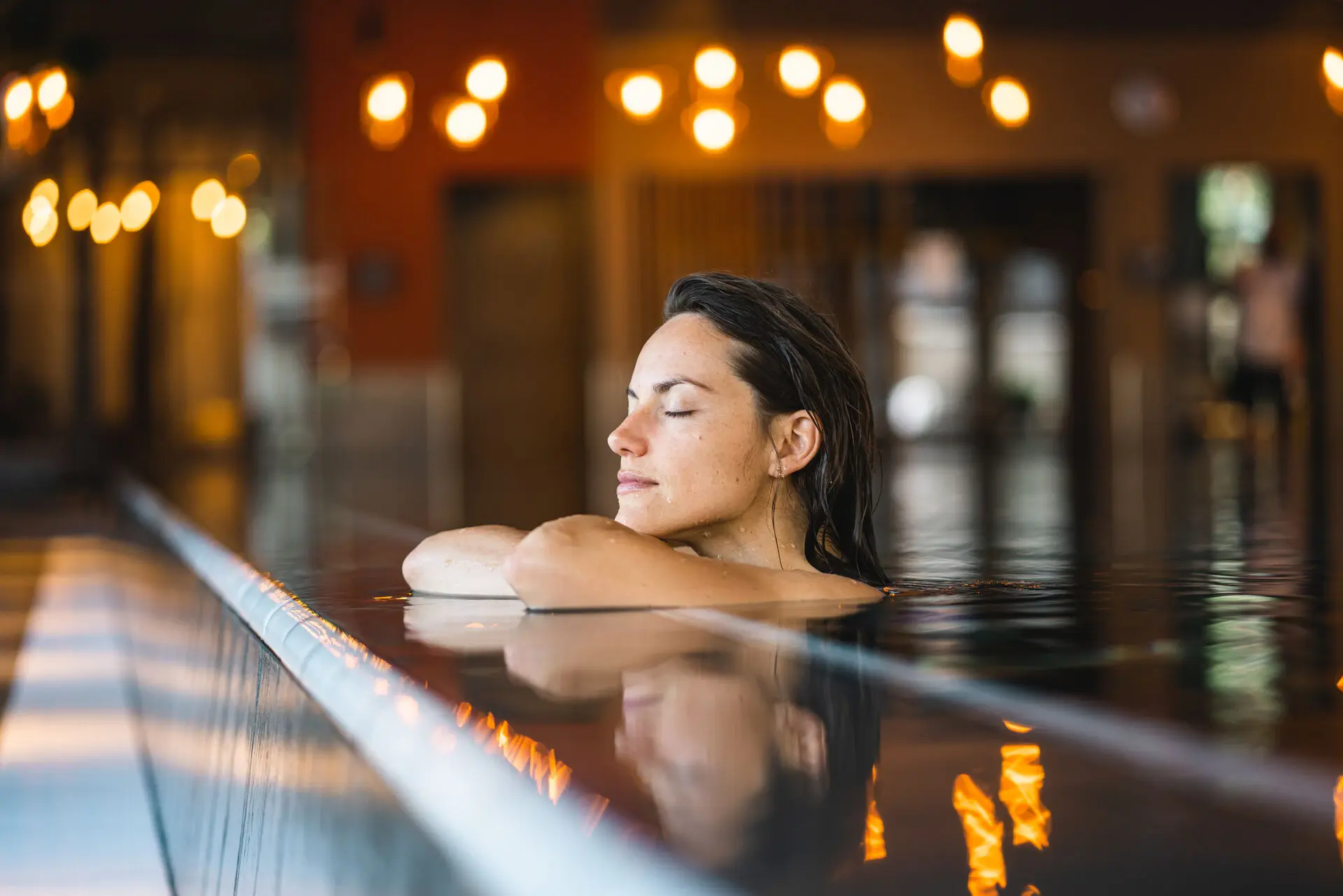 Eine Frau entspannt sich im warmen Wasser eines Indoor-Pools, mit geschlossenen Augen und einem friedlichen Gesichtsausdruck.